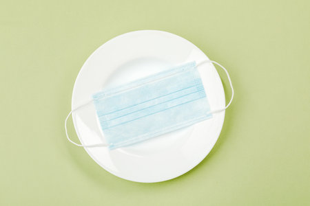 A medical mask on an empty plate with a fork and knife on a green background. The concept of nutrition and protection against the virusの写真素材