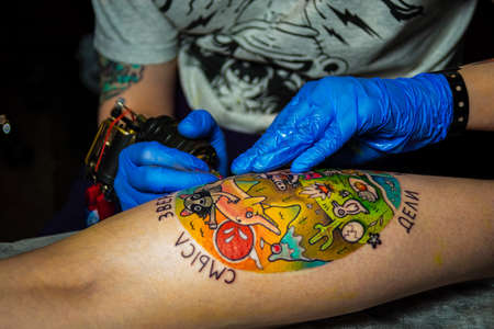 Tattoo artist holding machine in blue gloves. Close up creating picture on leg, abdomen, paper on client, blue lightのeditorial素材