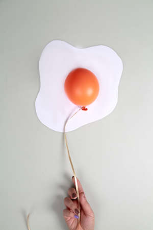 Orange balloon with white paper on grey background in shape of an egg. Food concept. Hand keep the balloon.の写真素材
