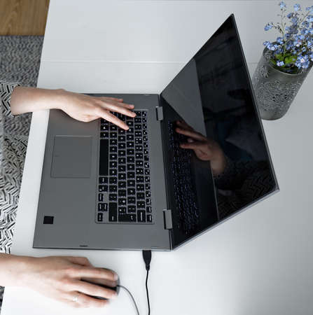 Female hand on grey laptop computer keyboard and black mouse on white desk.