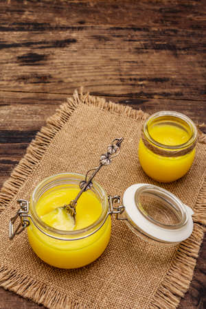 Pure or desi ghee (ghi), clarified melted butter. Healthy fats bulletproof diet concept or paleo style plan. Glass jars, silver spoon on vintage sackcloth. Wooden boards background, copy spaceの写真素材