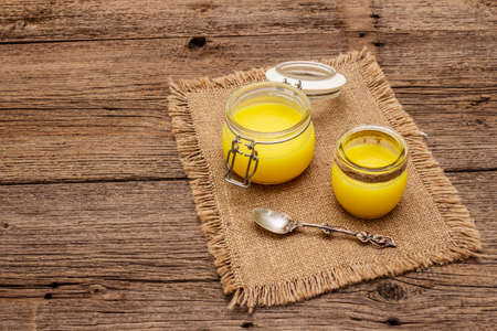 Pure or desi ghee (ghi), clarified melted butter. Healthy fats bulletproof diet concept or paleo style plan. Glass jars, silver spoon on vintage sackcloth. Wooden boards background, close upの写真素材