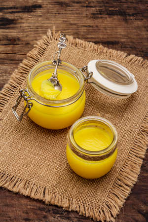 Pure or desi ghee (ghi), clarified melted butter. Healthy fats bulletproof diet concept or paleo style plan. Glass jars, silver spoon on vintage sackcloth. Wooden boards background, copy spaceの写真素材