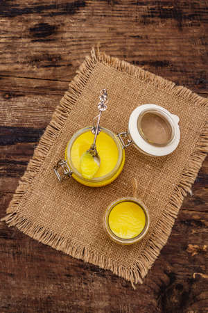 Pure or desi ghee (ghi), clarified melted butter. Healthy fats bulletproof diet concept or paleo style plan. Glass jars, silver spoon on vintage sackcloth. Wooden boards background, copy space, top viewの写真素材