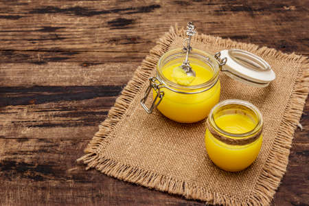Pure or desi ghee (ghi), clarified melted butter. Healthy fats bulletproof diet concept or paleo style plan. Glass jars, silver spoon on vintage sackcloth. Wooden boards background, copy spaceの写真素材