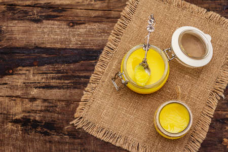 Pure or desi ghee (ghi), clarified melted butter. Healthy fats bulletproof diet concept or paleo style plan. Glass jars, silver spoon on vintage sackcloth. Wooden boards background, copy space, top viewの写真素材