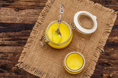 Pure or desi ghee (ghi), clarified melted butter. Healthy fats bulletproof diet concept or paleo style plan. Glass jars, silver spoon on vintage sackcloth. Wooden boards background, copy space, top viewの写真素材