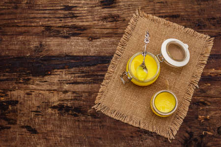 Pure or desi ghee (ghi), clarified melted butter. Healthy fats bulletproof diet concept or paleo style plan. Glass jars, silver spoon on vintage sackcloth. Wooden boards background, copy space, top viewの写真素材
