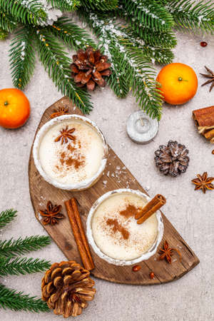 Eggnog with cinnamon and nutmeg for Christmas and winter holidays. Homemade beverage in glasses with spicy rim. Tangerines, candles, gift. Stone concrete background, top view, close upの写真素材