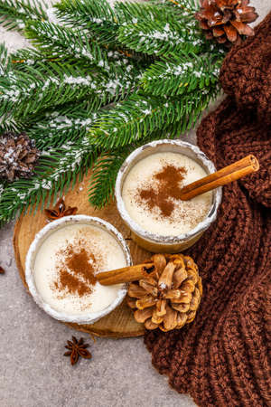 Homemade eggnog with cinnamon in glass. Typical Christmas dessert. Evergreen fir brunch, cones, cozy plaid, artificial snow. Stone concrete background, top view, close upの写真素材
