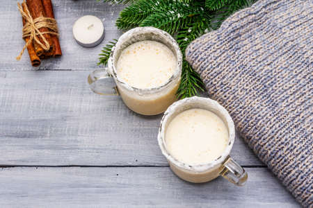 Eggnog cocktail in two glasses mug arranged with christmas decoration on light wooden table. Evergreen fir tree branch, gift, cozy jersey, artificial snow, copy space, top view, close upの写真素材