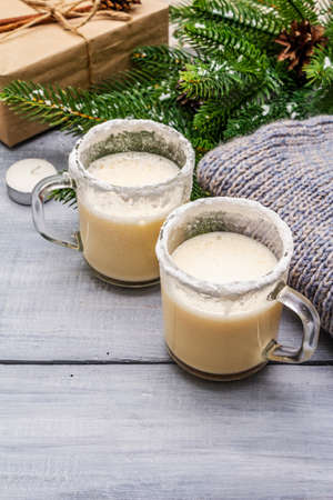 Eggnog cocktail in two glasses mug arranged with christmas decoration on light wooden table. Evergreen fir tree branch, gift, cozy jersey, artificial snow, copy space, close upの写真素材