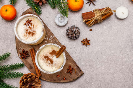 Eggnog with cinnamon and nutmeg for Christmas and winter holidays. Homemade beverage in glasses with spicy rim. Tangerines, candles, gift. Stone concrete background, copy space, top viewの写真素材