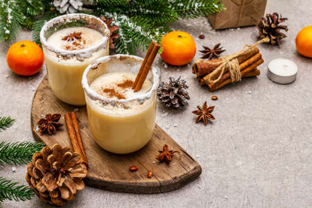 Eggnog with cinnamon and nutmeg for Christmas and winter holidays. Homemade beverage in glasses with spicy rim. Tangerines, candles, gift. Stone concrete background, copy space, close upの写真素材