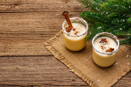 Eggnog cocktail in two glasses arranged with christmas decoration on old wooden table. Evergreen fir tree branch, artificial snow, sackcloth napkin, copy spaceの写真素材