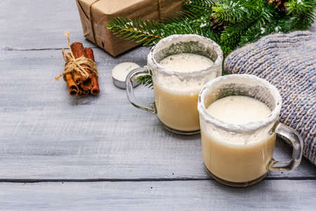 Eggnog cocktail in two glasses mug arranged with christmas decoration on light wooden table. Evergreen fir tree branch, gift, cozy jersey, artificial snow, copy spaceの写真素材