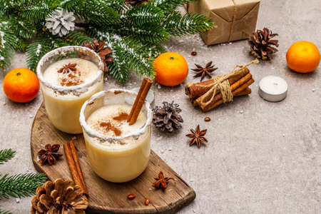 Eggnog with cinnamon and nutmeg for Christmas and winter holidays. Homemade beverage in glasses with spicy rim. Tangerines, candles, gift. Stone concrete background, close upの写真素材