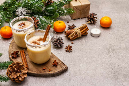 Eggnog with cinnamon and nutmeg for Christmas and winter holidays. Homemade beverage in glasses with spicy rim. Tangerines, candles, gift. Stone concrete background, copy spaceの写真素材
