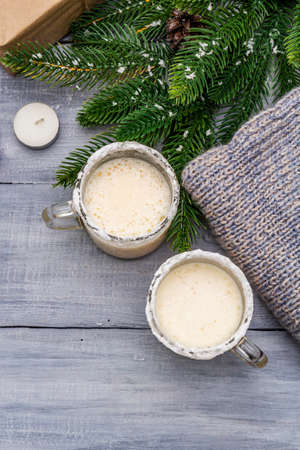 Eggnog cocktail in two glasses mug arranged with christmas decoration on light wooden table. Evergreen fir tree branch, gift, cozy jersey, artificial snow, copy space, top view, close upの写真素材