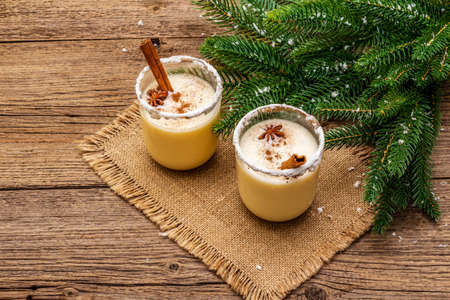 Eggnog cocktail in two glasses arranged with christmas decoration on old wooden table. Evergreen fir tree branch, artificial snow, sackcloth napkin, close upの写真素材