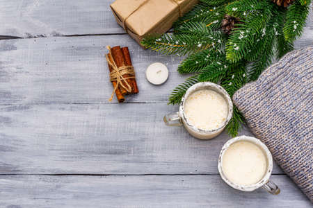 Eggnog cocktail in two glasses mug arranged with christmas decoration on light wooden table. Evergreen fir tree branch, gift, cozy jersey, artificial snow, copy space, top viewの写真素材