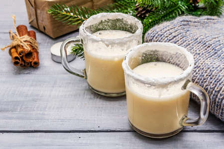 Eggnog cocktail in two glasses mug arranged with christmas decoration on light wooden table. Evergreen fir tree branch, gift, cozy jersey, artificial snow, copy space, close upの写真素材