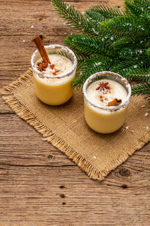 Eggnog cocktail in two glasses arranged with christmas decoration on old wooden table. Evergreen fir tree branch, artificial snow, sackcloth napkin, copy spaceの写真素材