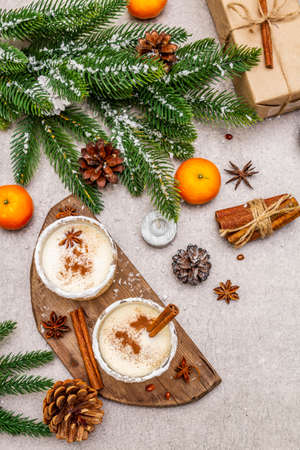 Eggnog with cinnamon and nutmeg for Christmas and winter holidays. Homemade beverage in glasses with spicy rim. Tangerines, candles, gift. Stone concrete background, top view, close upの写真素材