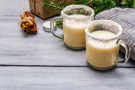 Eggnog cocktail in two glasses mug arranged with christmas decoration on light wooden table. Evergreen fir tree branch, gift, cozy jersey, artificial snow, copy spaceの写真素材