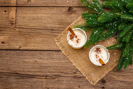 Eggnog cocktail in two glasses arranged with christmas decoration on old wooden table. Evergreen fir tree branch, artificial snow, sackcloth napkin, copy space, top viewの写真素材