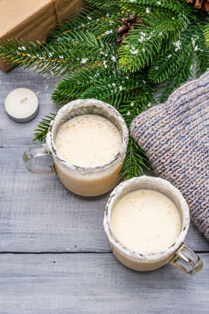 Eggnog cocktail in two glasses mug arranged with christmas decoration on light wooden table. Evergreen fir tree branch, gift, cozy jersey, artificial snow, copy space, top view, close upの写真素材