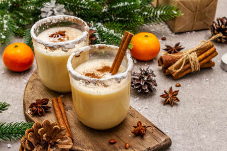 Eggnog with cinnamon and nutmeg for Christmas and winter holidays. Homemade beverage in glasses with spicy rim. Tangerines, candles, gift. Stone concrete background, close upの写真素材