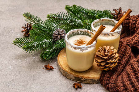 Homemade eggnog with cinnamon in glass. Typical Christmas dessert. Evergreen fir brunch, cones, cozy plaid, artificial snow. Stone concrete backgroundの写真素材