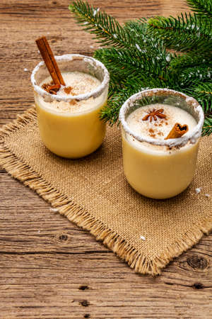 Eggnog cocktail in two glasses arranged with christmas decoration on old wooden table. Evergreen fir tree branch, artificial snow, sackcloth napkin, copy space, close upの写真素材