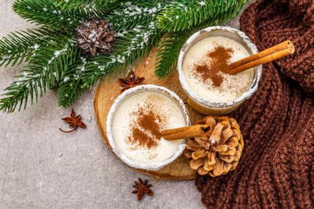 Homemade eggnog with cinnamon in glass. Typical Christmas dessert. Evergreen fir brunch, cones, cozy plaid, artificial snow. Stone concrete background, top view, close upの写真素材