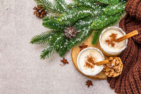 Homemade eggnog with cinnamon in glass. Typical Christmas dessert. Evergreen fir brunch, cones, cozy plaid, artificial snow. Stone concrete background, copy space, top viewの写真素材