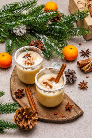 Eggnog with cinnamon and nutmeg for Christmas and winter holidays. Homemade beverage in glasses with spicy rim. Tangerines, candles, gift. Stone concrete background, close upの写真素材