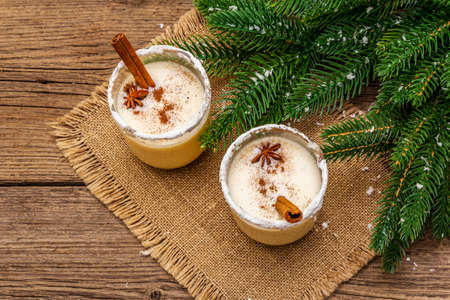 Eggnog cocktail in two glasses arranged with christmas decoration on old wooden table. Evergreen fir tree branch, artificial snow, sackcloth napkin, top view, close upの写真素材