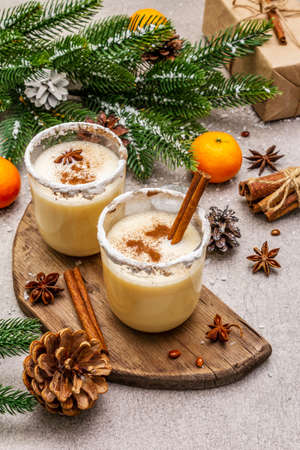 Eggnog with cinnamon and nutmeg for Christmas and winter holidays. Homemade beverage in glasses with spicy rim. Tangerines, candles, gift. Stone concrete background, close upの写真素材