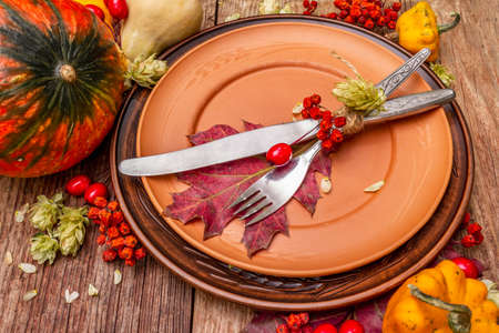 Autumn and Thanksgiving dinner place setting. Harvest of vegetables and berries, colorful leaves, hops, ceramic plates, fork and knife. Fall cutlery wooden background, close upの写真素材