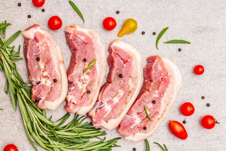 Raw pork belly with rind, peritoneum meat. Fresh tomato cherry, rosemary, spice. Ingredient for cooking healthy meal, animal proteins. Light stone concrete background top view close upの写真素材