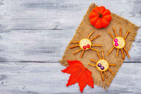 Funny kids food. Edible spiders from cookies, small candies and sweet sesame sticks. Halloween concept with autumn leaves, pumpkin. Vintage sackcloth, wooden boards background, copy space, top viewの写真素材