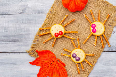 Funny kids food. Edible spiders from cookies, small candies and sweet sesame sticks. Halloween concept with autumn leaves, pumpkin. Wooden boards background, copy space top view close upの写真素材
