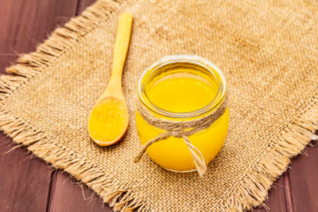 Pure or desi ghee (ghi), clarified melted butter. Healthy fats bulletproof diet concept or paleo style plan. Glass jar, wooden spoon on vintage sackcloth. Wooden boards background, copy spaceの写真素材