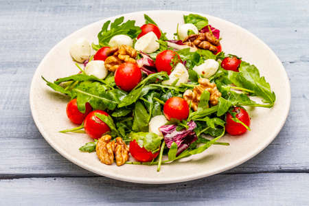 Fresh salad with arugula, radicchio, tomato cherry, baby mozzarella, walnuts. Olive oil, sea salt, ground black pepper. Healthy eating food. Wooden boards background close upの写真素材