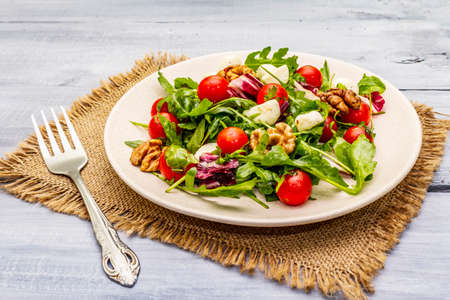 Fresh salad with arugula, radicchio, tomato cherry, baby mozzarella, walnuts. Olive oil, sea salt, ground black pepper. Healthy eating food. Vintage sackcloth, wooden boards background, close upの写真素材