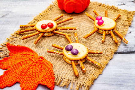 Funny kids food. Edible spiders from cookies, small candies and sweet sesame sticks. Halloween concept with autumn leaves, pumpkin. Vintage sackcloth, wooden boards background, close upの写真素材