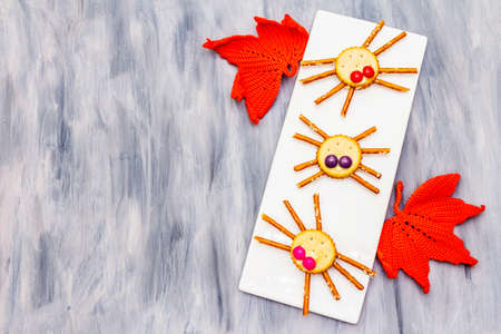 Funny kids food. Edible spiders from cookies, small candies and sweet sesame sticks. Halloween concept with autumn leaves. Ceramic plate, wooden painted abstract background copy space top viewの写真素材