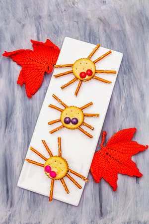 Funny kids food. Edible spiders from cookies, small candies and sweet sesame sticks. Halloween concept with autumn leaves. Ceramic plate, wooden painted abstract background, top view, close upの写真素材