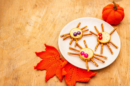 Funny kids food. Edible spiders from cookies, small candies and sweet sesame sticks. Halloween concept with autumn leaves, pumpkin. Ceramic plate, wooden background, copy spaceの写真素材